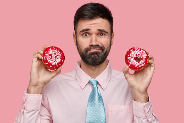 Free photo headshot of puzzled unshaven man dressed formally, holds two doughnuts in both hands