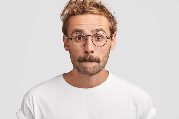 Free photo headshot of puzzled young man presses lips and looks in bewilderment, feels puzzlement, has hesitant expression, contemplates about something