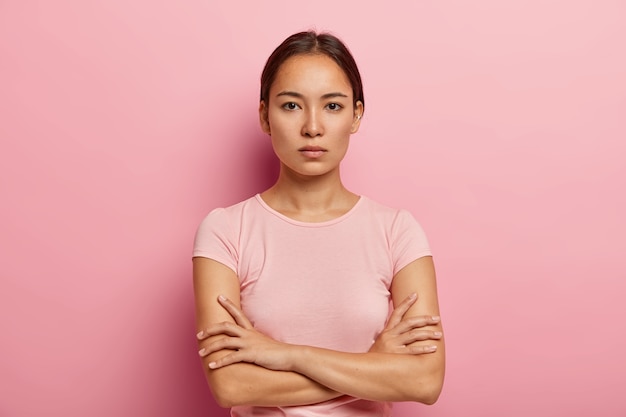 Headshot of serious korean woman looks with calm face expression, keeps arms folded, has healthy fresh skin, wears rosy t shirt, stands indoor. Beautiful Asian girl has confident gaze 