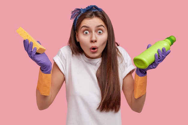 Headshot of surprised Caucasian young woman looks in amazement, wears casual clothes, holds rag and bottle of detergent