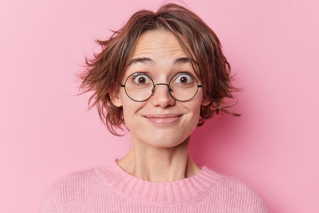 Free Photo headshot of surprised happy woman stares through glasses cannot believe in shocking relevation has trendy bob hairstyle wears casual jumper isolated over pink background hears something pleasant