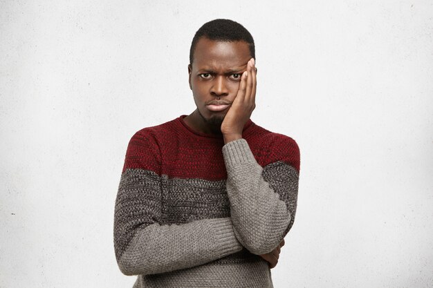 Headshot of unhappy frustrated young black male having puzzled expression, holding hand on his cheek and keeping arms folded. Sad African American man dressed in sweater feeling bored or depressed