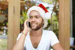 Free photo headshot of young unshaven man dressed in white t-shirt and red santa claus hat looking happy while speaking on mobile phone with his girlfriend, listening to her warm congratulations on christmas