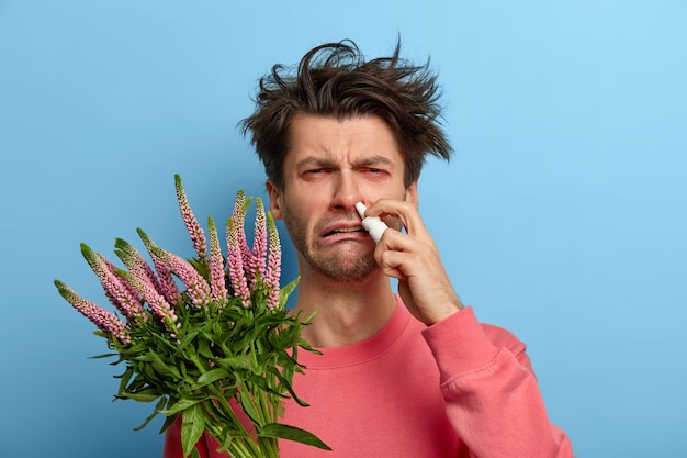 Health care and allergy symptoms concept. Dissatisfied man cures allergic rhinitis with nasal drops, has sick expression caused by trigger, annoyed red eyes, poses indoor, being hypersensitive