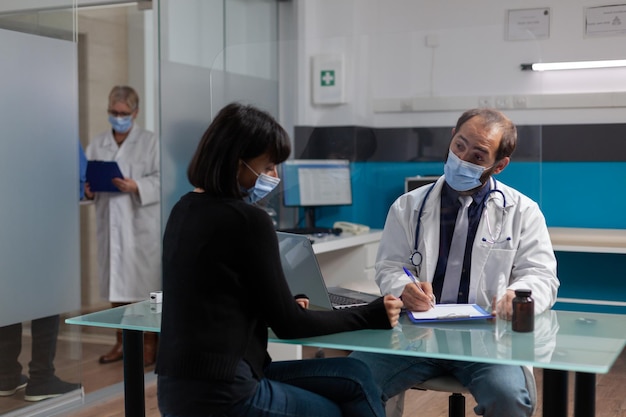 Health physician taking notes at checkup consultation in cabinet, doing examination with patient to cure illness. Doctor writing diagnosis and treatment on paper during covid 19 pandemic.