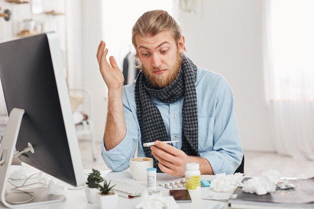 Health problems concept. Astonished young bearded office worker has a bad cold, flu, looks at thermometer with bugged eyes after measuring temperature of body. Sick manager against office background