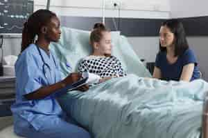 Free photo healthcare pediatric clinic nurse taking notes about successful recovery while in patient room. mother being attentive to nurse speaking about little girl health hazards while in recovery ward.