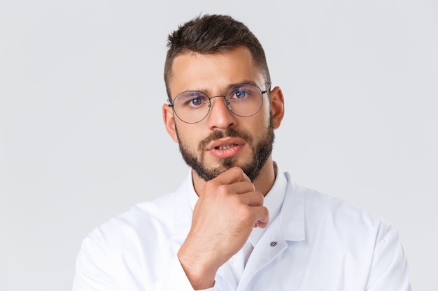 Healthcare workers, coronavirus, covid-19 pandemic concept. Close-up of serious-looking professional doctor in glasses and white coat, touch chin thoughtful, thinking, making decision on patient