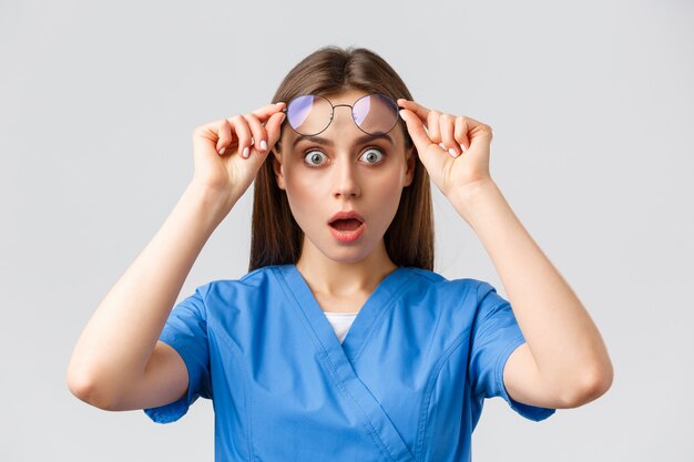 Healthcare workers, medicine, insurance and covid-19 pandemic concept. Surprised or shocked female nurse, doctor in blue scrubs, open mouth and take-off glasses astonished