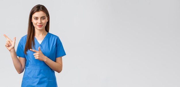 Healthcare workers prevent virus insurance and medicine concept Smiling attractive female doctor or nurse in blue scrubs pointing fingers left to show patients advertisement important info