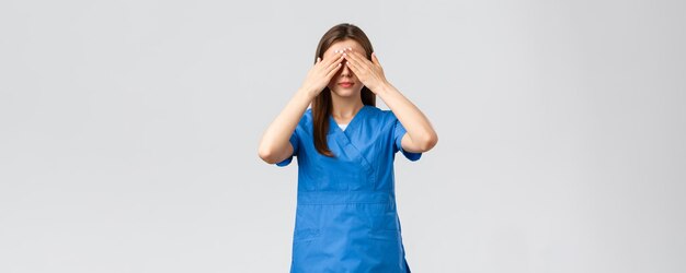 Healthcare workers prevent virus insurance and medicine concept Young woman in blue scrubs doctor or nurse close eyes with hands blindfolded medicine standing grey background