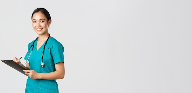 Healthcare workers, preventing virus, quarantine campaign concept. Smiling friendly female doctor, nurse in scrubs writing dorn results analysis at clipboard, standing white background.