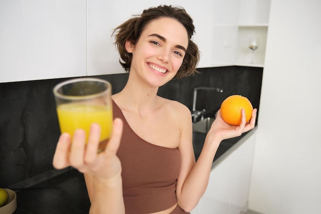 Free photo healthy body and wellbeing smiling fitness girl giving you glass of fresh juice holding an orange an