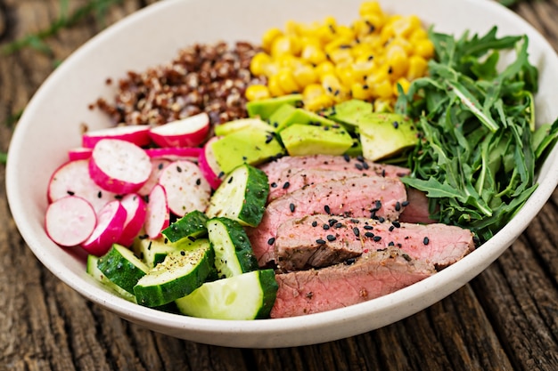 Free Photo healthy dinner. buddha bowl lunch with grilled beef steak and quinoa, corn, avocado, cucumber and arugula on wooden table. meat salad.