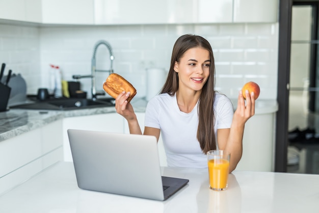 Healthy food concept. Hard choice. Yound sporty woman is choosing between healthy food and sweets while standing on light kitchen.