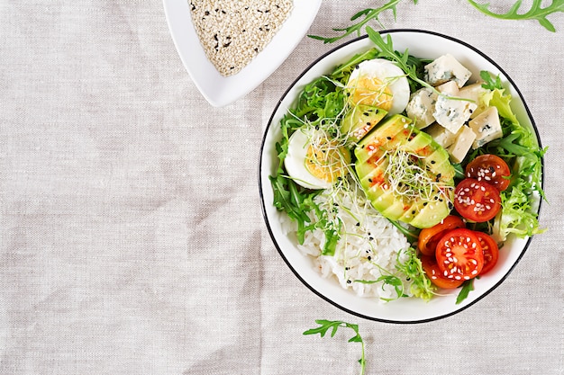 Healthy green vegetarian buddha bowl lunch with eggs, rice, tomato, avocado and blue cheese on table.