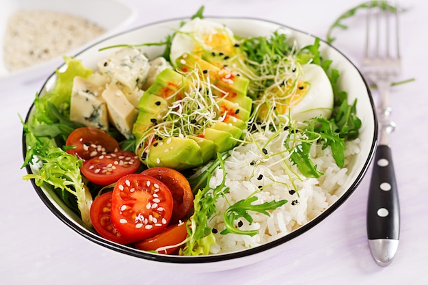 Healthy green vegetarian buddha bowl lunch with eggs, rice, tomato, avocado and blue cheese on table.