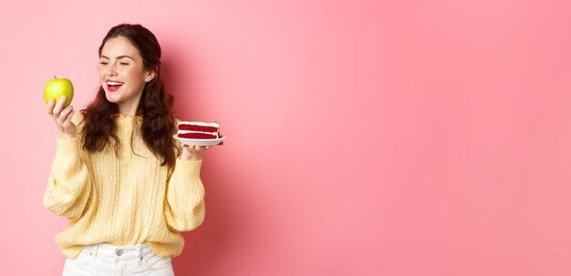 Free photo healthy lifestyle concept young brunette woman holding green apple full of vitamins and cake with lo