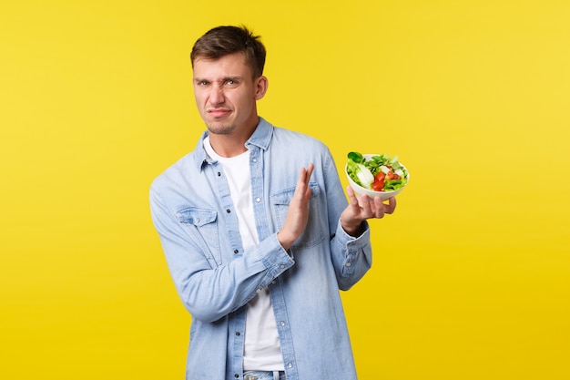 Free photo healthy lifestyle, people and food concept. disgusted and disappointed blond man grimacing from aversion and showing refusal to bowl with salad, dislike staying on diet, yellow background.