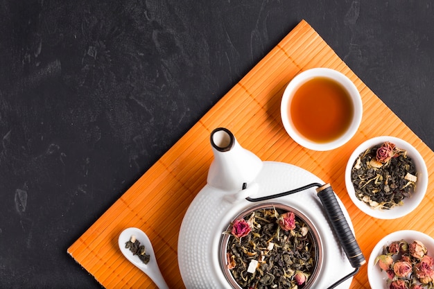 Healthy organic dried tea herb and teapot on orange placemat over black background