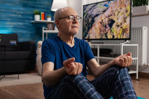 Healthy senior man sitting comfortable in lotus position on yoga mat with closed eyes