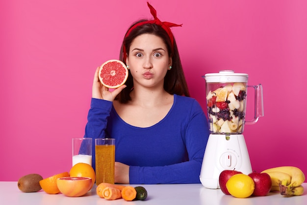 Healthy vegeterian girl openes her eyes widely, blows her mouth, looking directly at camera, holding fruit in one hand. Different fruit is on table and in blender, cooking delicious smoothie.