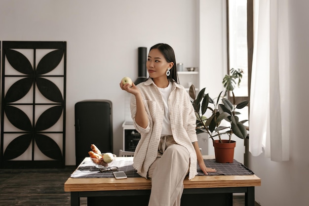 Free photo healthy young brunette asian woman in pants and beige cardigan sits on wooden table on kitchen smiles and holds fresh tasty apple