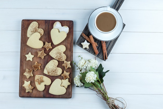 Free photo heart-shaped and star cookies on a wooden cutting board with cup of coffee, flowers, cinnamon flat lay on a white wooden board background