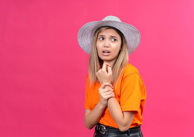 A helpless pretty young woman in an orange t-shirt wearing sunhat holding her painful wrist while looking side on a pink wall