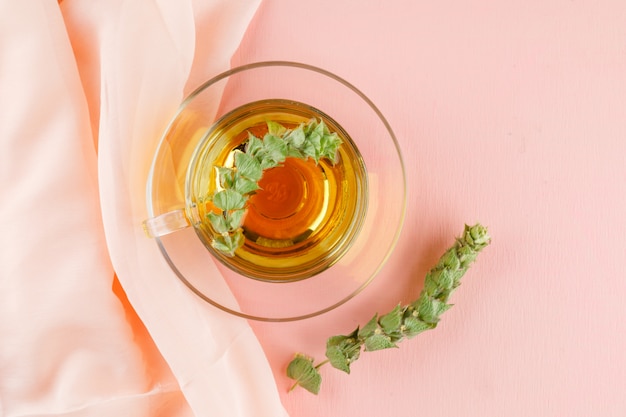 Herbal tea with herbs in a glass cup on pink and textile, flat lay.