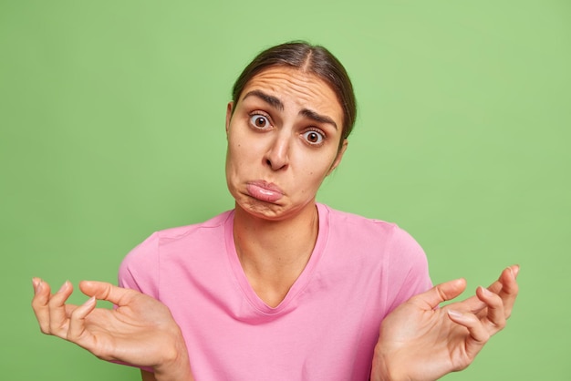 Free photo hesitant clueless unaware young woman spreads palms looks with sad doubtful expression purses lower lip poses against bright green background. discontent questioned female model shrugs shoulders