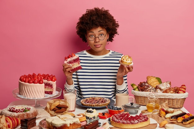 Free photo hesitant dark skinned curly woman feels doubt which piece of cake to choose, has temptation to eat junk food, poses at big festive table with desserts against pink background