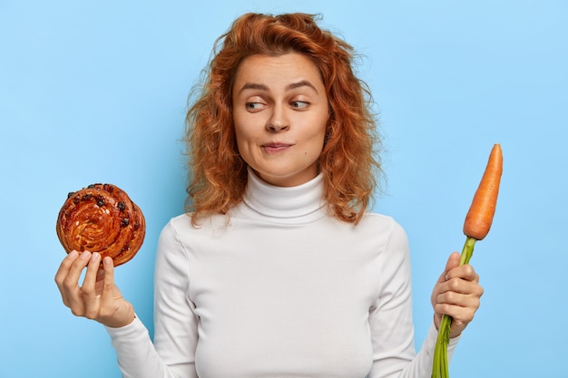 Free photo hesitant foxy girl has doubt between junk food and fresh vegetable, keeps to diet, holds baked delicious roll and carrot, feels temptation, wears casual outfit, models indoor. nutrition concept