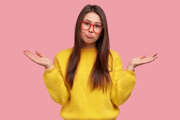 Free Photo hesitant indecisive young woman purses lower lip, spreads palms with clueless expression, dressed in casual warm yellow jumper, being unaware