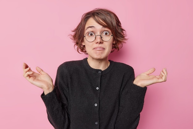 Free Photo hesitant puzzled young european woman with short hairstyle spreads palms looks clueless cannot mae decision wears round spectacles and black jumper isolated over pink background. so what to do