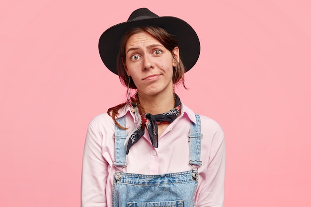 Free photo hesitant successful woman gardener in denim overalls, bandana on neck, hat on head, has puzzled clueless expression