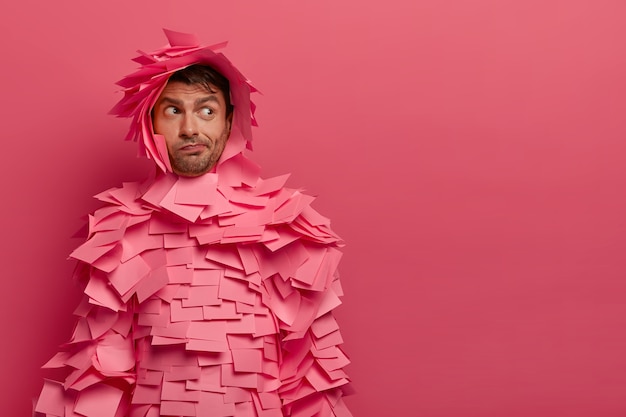 Free photo hesitant unshaven young man looks aside, wears paper costume, uses office sticky notes, thinks about something, poses against pink wall, copy space for your advertisement or promotion.