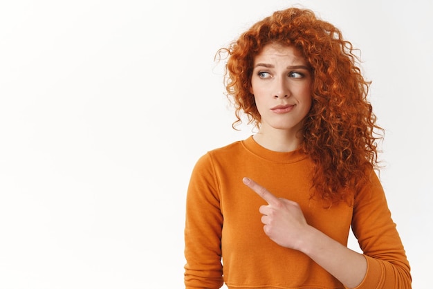 Hesitant worried cute modest redhead curlyhaired female looking pointing left dissatisfied and doubtful have concern about bad uncertain choice standing white background disappointed