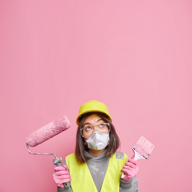 Free photo hesitant young professional asian female decorator holds painting tools thinks about house redecoration focused above wears protective helmet respirator and safety glasses isolated on pink wall