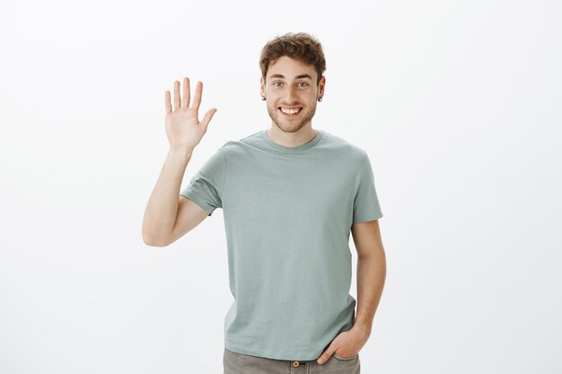 Hi nice to meet you. Portrait of handsome outgoing european guy in casual t-shirt raising hand and waving palm in hello gesture