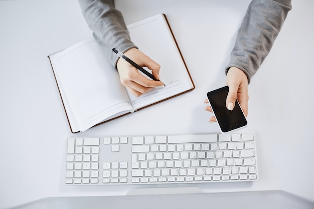 Free Photo high angle shot of busy hardworking woman holding smartphone. cropped shot of woman hands making notes in planner and sitting near keyboard and computer, being at work and working with teammates