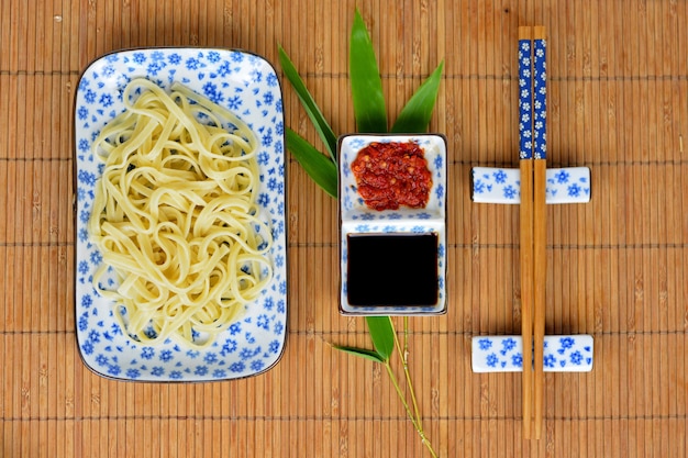 High angle shot of noodles and sauces in white plates and chopsticks on a bamboo table cover