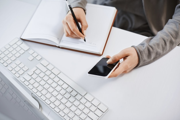 Free Photo high angle shot of woman hands working with gadgets. cropped shot of modern female holding smartphone while writing plan in notebook, sitting near keyboard and computer, having tough time at office