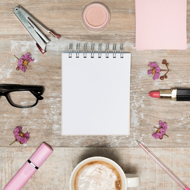 Free photo high angle view of blank white notepad surrounded by cosmetic products; coffee cup; flowers and eyeglass arranged on wooden desk