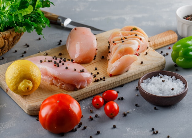 High angle view chicken breasts on cutting board with lemon, tomatoes, salt on gray surface