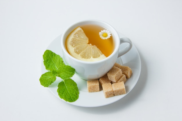 High angle view a cup of chamomile tea with lemon, mint leaves, sugar on white surface. horizontal