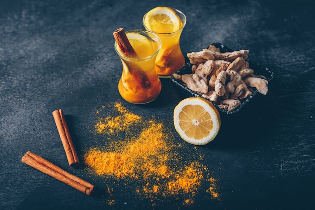 Free Photo high angle view ginger powder in tea cups and a bowl with lemon, ginger and dry cinnamon on dark textured background. horizontal