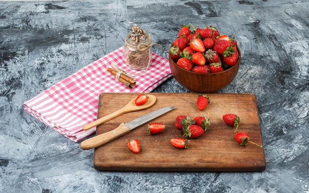 High angle view a glass jar and cinnamon on red gingham tablecloth with kitchen utensils and a bowl of strawberries on dark blue marble surface. horizontal