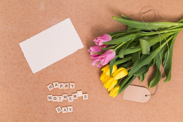 Free photo high angle view of happy mother's day text; blank paper and yellow; pink tulip flowers above brown backdrop