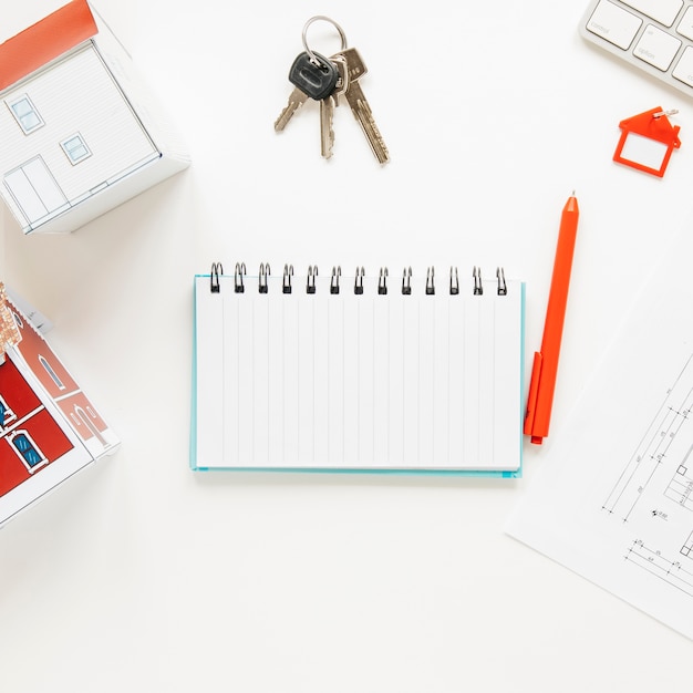 High angle view of house model near spiral notepad with keys and pen over white background